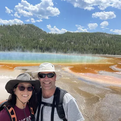 Irene and her husband in sun hats beside a brightly colored hot spring, with a forested ridge behind.
