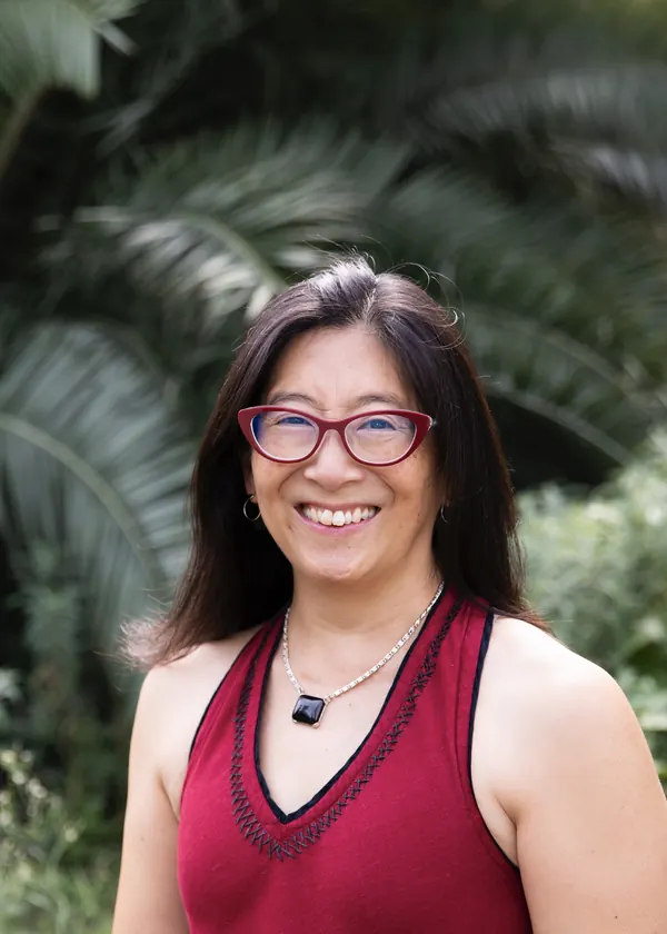 Irene Salter, smiling, in red glasses and a crimson top, with palm fronds behind her.